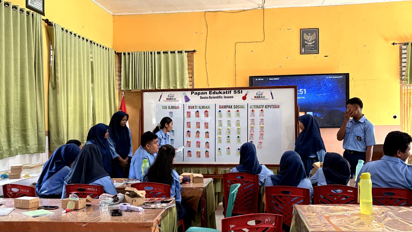 Siswi SMAN 2 Siak Hulu berseragam biru memegang poster bergambar pohon kelapa sawit sedang menjelaskan kepada tiga dosen UIR berjilbab yang menyimak dan mencatat saat kegiatan Deep Learning SSI di sekolah