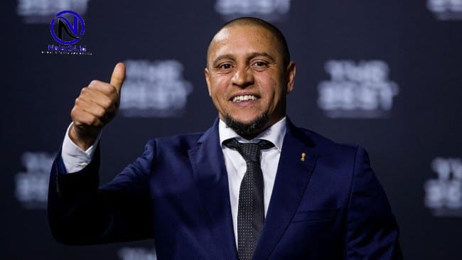 Roberto Carlos memakai jas biru dongker mengacungkan jempol di karpet merah
