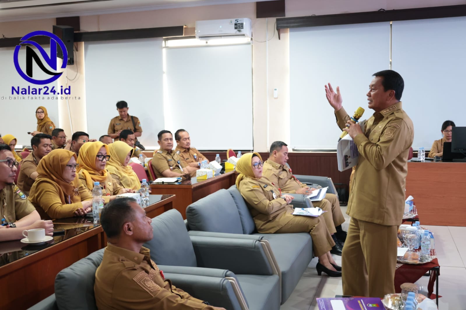 Seorang pejabat memakai seragam coklat berdiri sambil memegang mikrofon dan berbicara di depan para peserta rapat di dalam aula