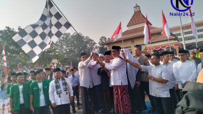 Seorang pejabat memakai baju putih dan sarung batik memegang bendera kotak hitam putih bersama rombongan di atas panggung