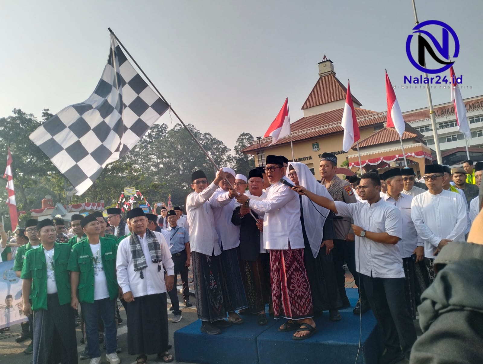 Seorang pejabat memakai baju putih dan sarung batik memegang bendera kotak hitam putih bersama rombongan di atas panggung