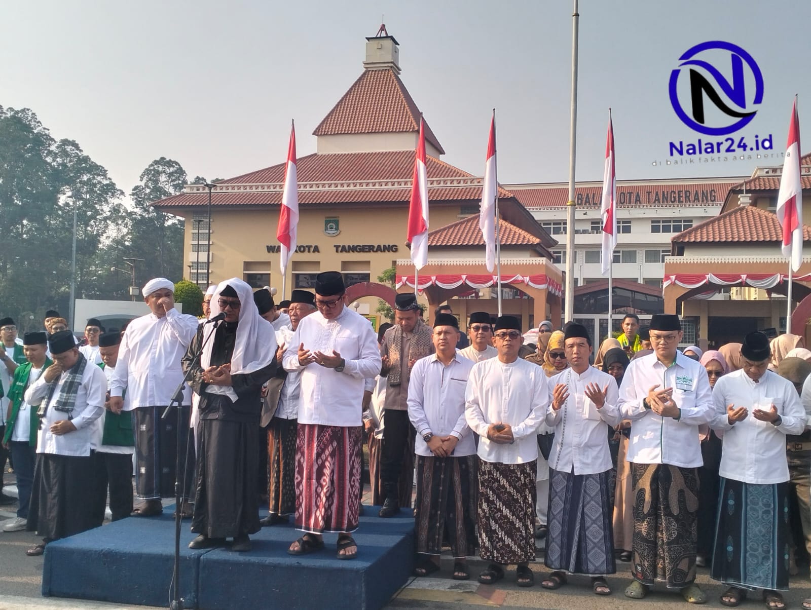 Sejumlah pria memakai baju koko putih dan sarung berdiri dengan tangan terangkat berdoa di depan gedung Balai Kota