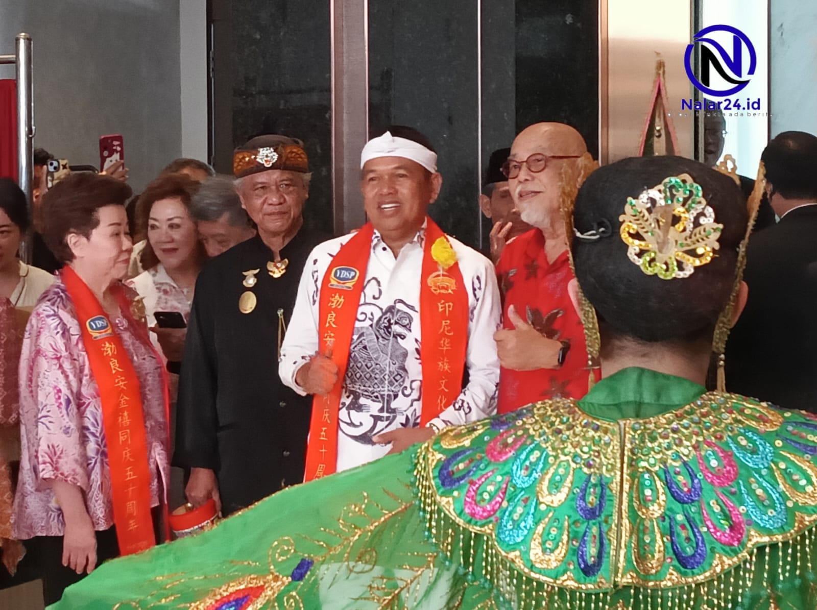 Seorang pria memakai baju putih motif batik dan selempang merah berdiri di tengah sambil tersenyum melihat penari memakai baju hijau bordir emas