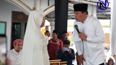 Seorang pejabat pria memakai baju koko putih dan peci hitam memegang mikrofon dan kertas sambil berbicara dengan seorang wanita berhijab putih