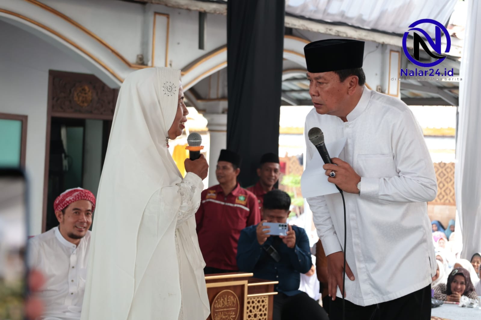 Seorang pejabat pria memakai baju koko putih dan peci hitam memegang mikrofon dan kertas sambil berbicara dengan seorang wanita berhijab putih