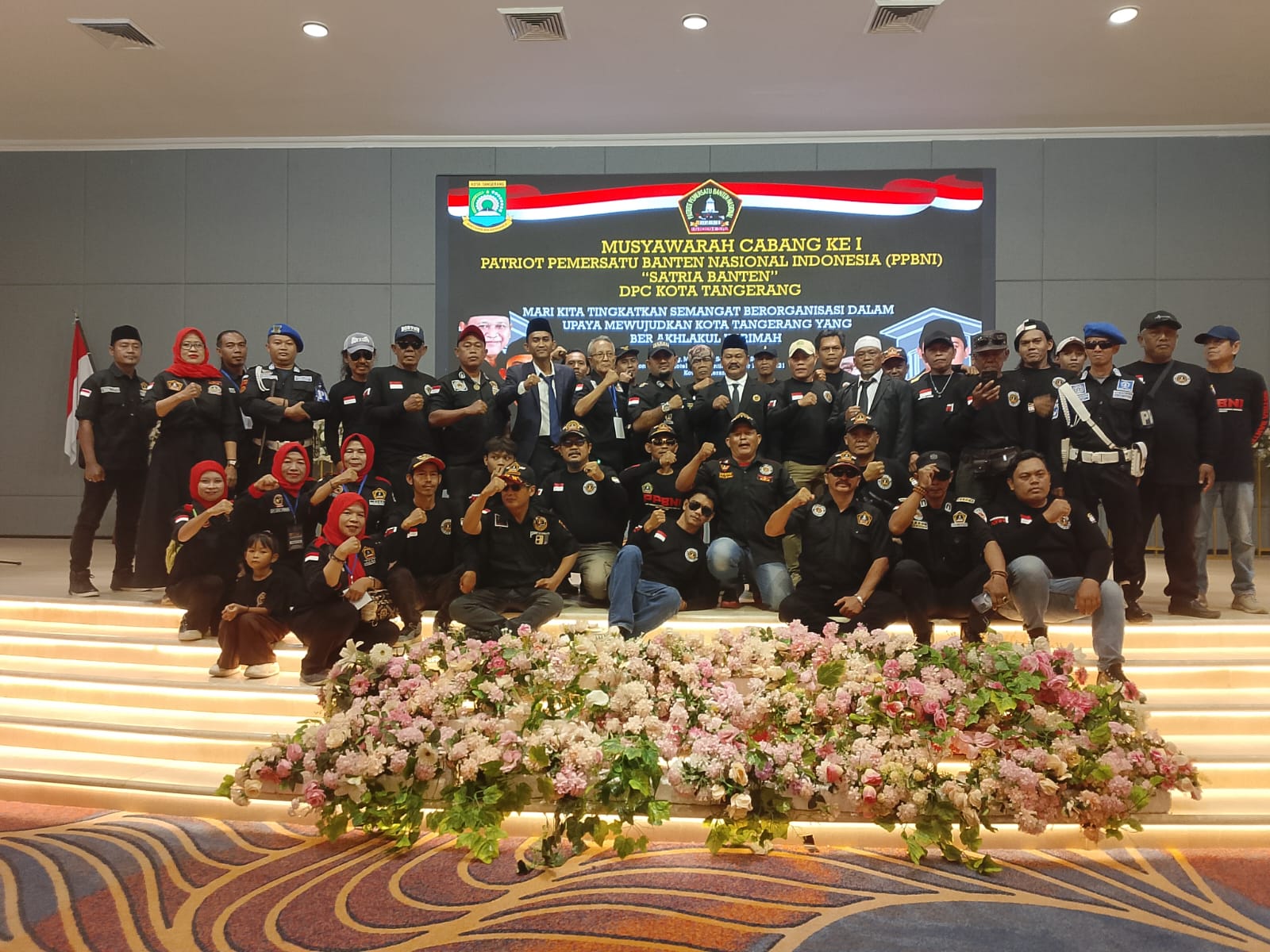 Foto bersama puluhan anggota PPBNI Satria Banten DPC Kota Tangerang di atas panggung setelah Musyawarah Cabang I. Peserta memakai seragam hitam dengan latar layar bertuliskan Muscab I PPBNI dan hiasan bunga,